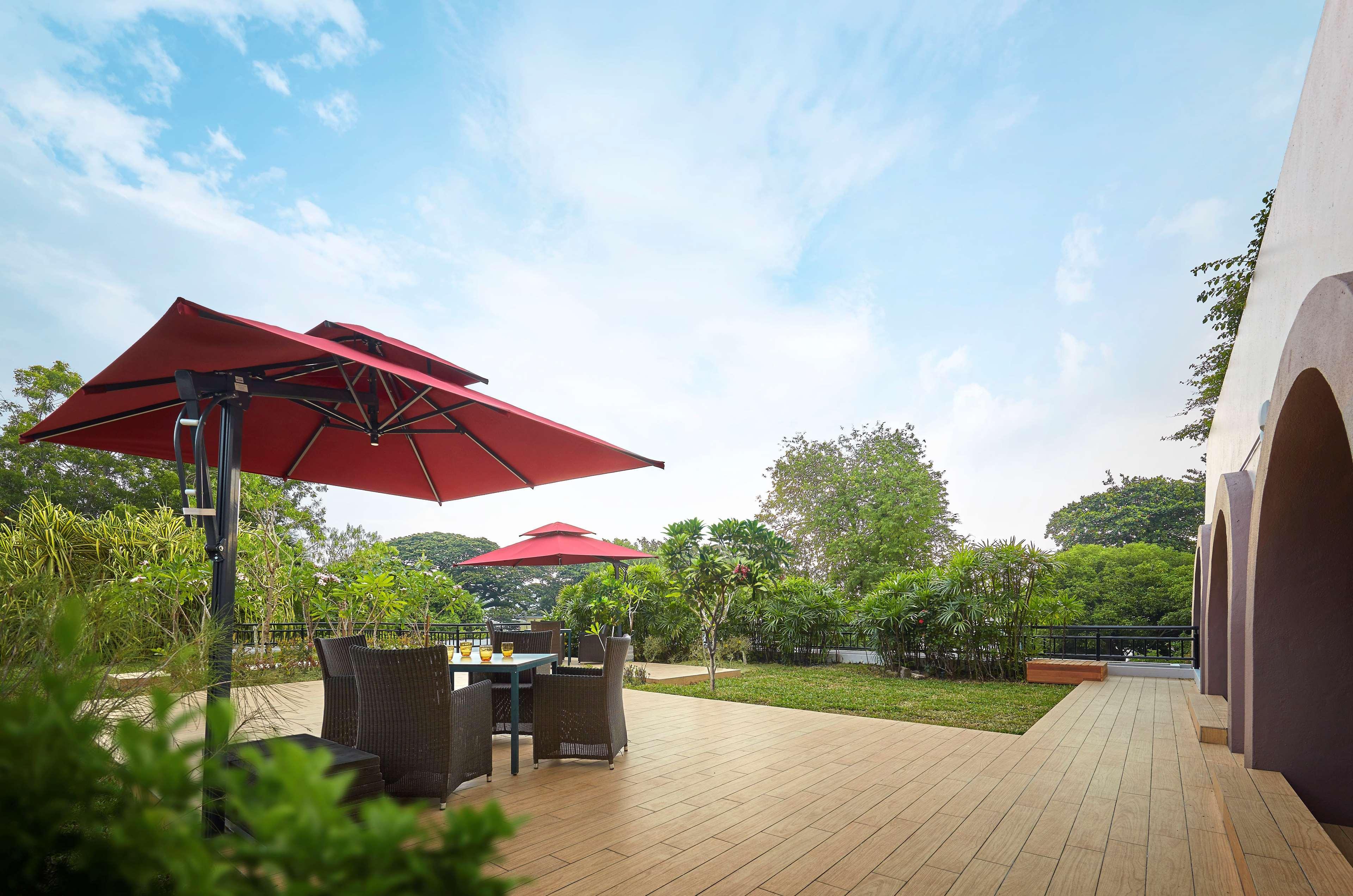 Parkroyal Penang Resort Batu Ferringhi Exterior photo The photo shows an outdoor patio area with a wooden deck. There are two red umbrellas providing shade over a small dining table with dark chairs. Surrounding the area are lush green plants and trees, contributing to a serene atmosphere. The sky above