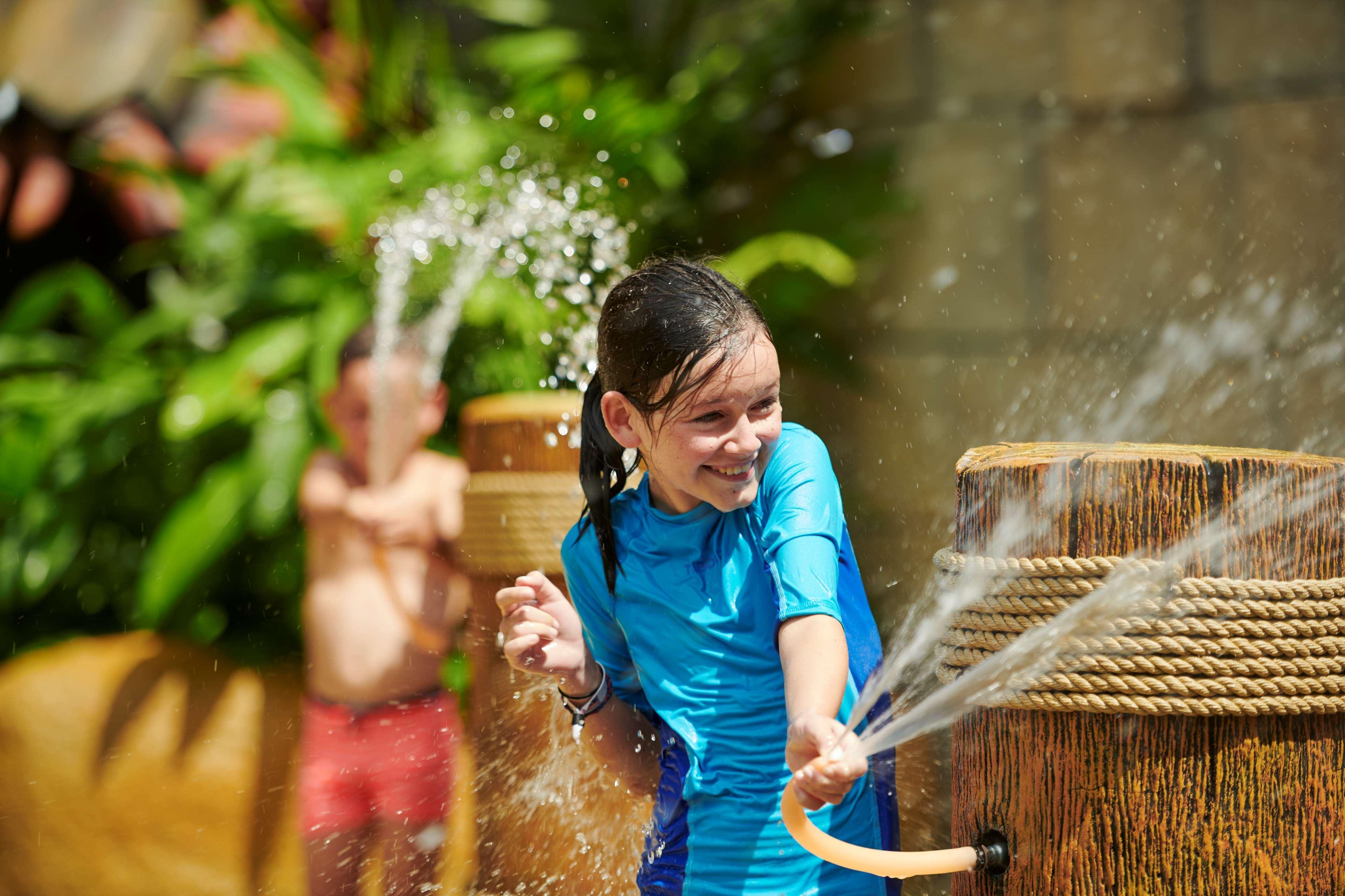 Parkroyal Penang Resort Batu Ferringhi Exterior photo The photo depicts a joyful scene at a water park or a similar environment. In the foreground, a girl wearing a blue swimsuit is smiling as she interacts with a water feature that sprays water. She appears to be having a lot of fun. In the background,