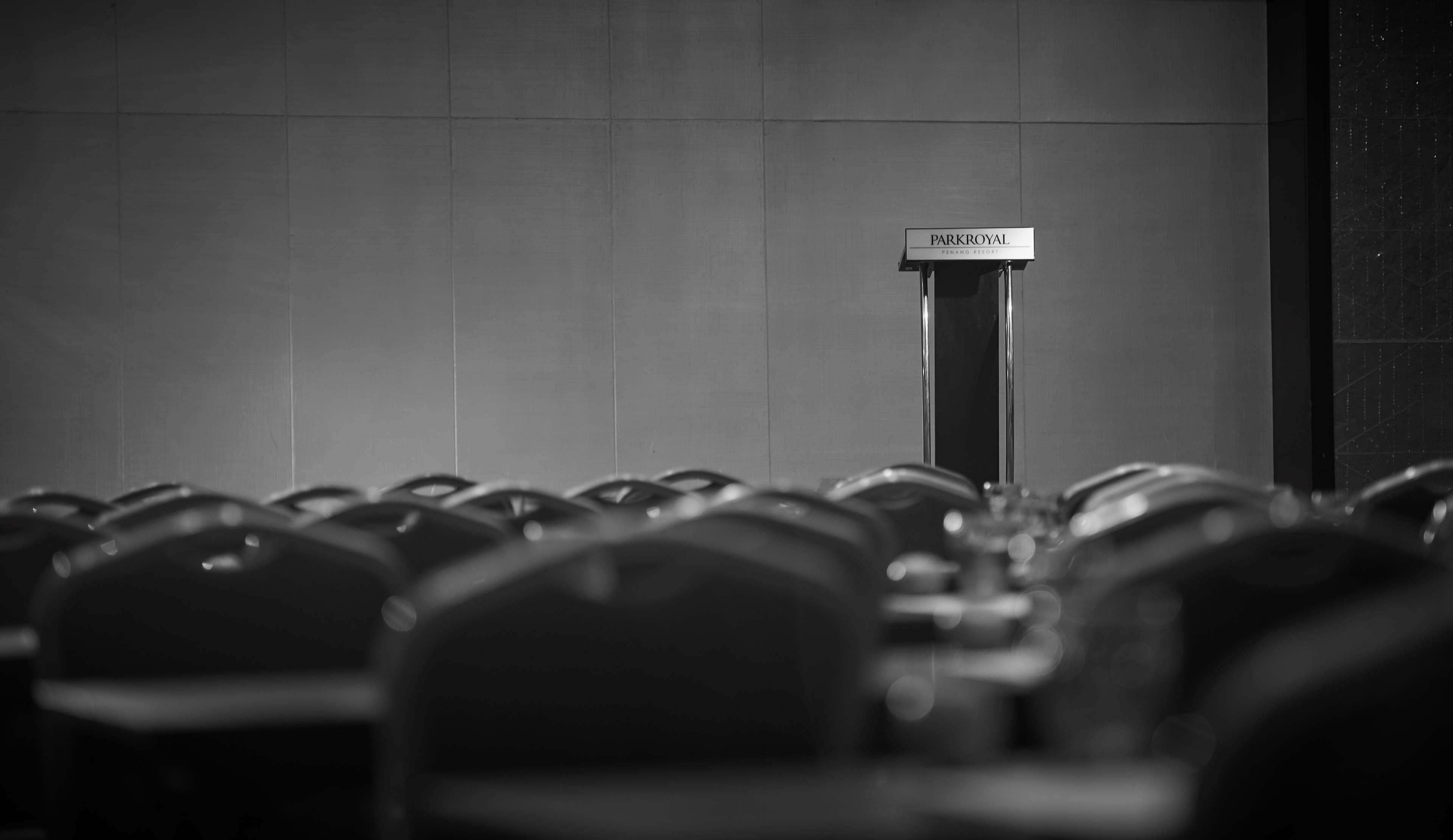 Parkroyal Penang Resort Batu Ferringhi Exterior photo The photo depicts a podium or lectern at the front of a conference room or hall. The podium is prominently placed against a plain background, hinting at a formal setting. In the foreground, there are several rows of empty chairs arranged neatly, indi