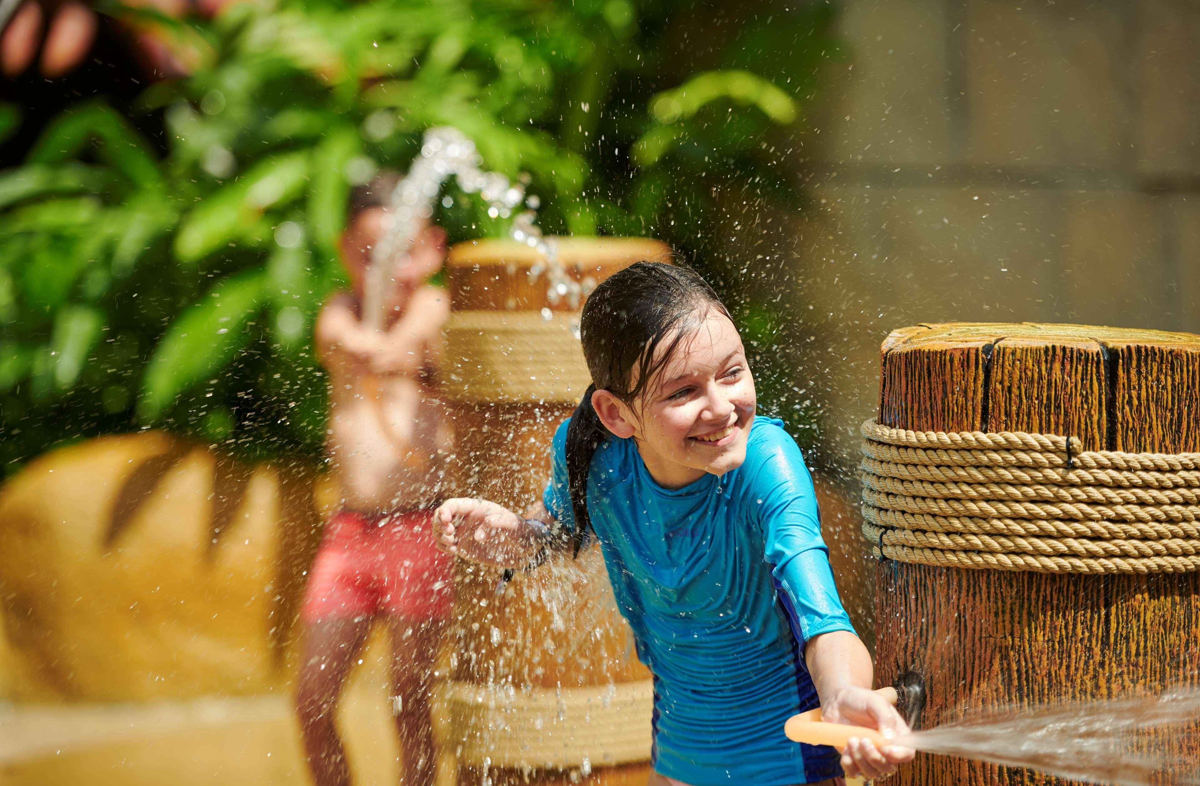 Parkroyal Penang Resort Batu Ferringhi Exterior photo The photo depicts a playful scene at a water park or pool area. In the foreground, a young girl is smiling and enjoying herself while interacting with water, wearing a blue swimsuit. Behind her, another child is seen in the background, seemingly play