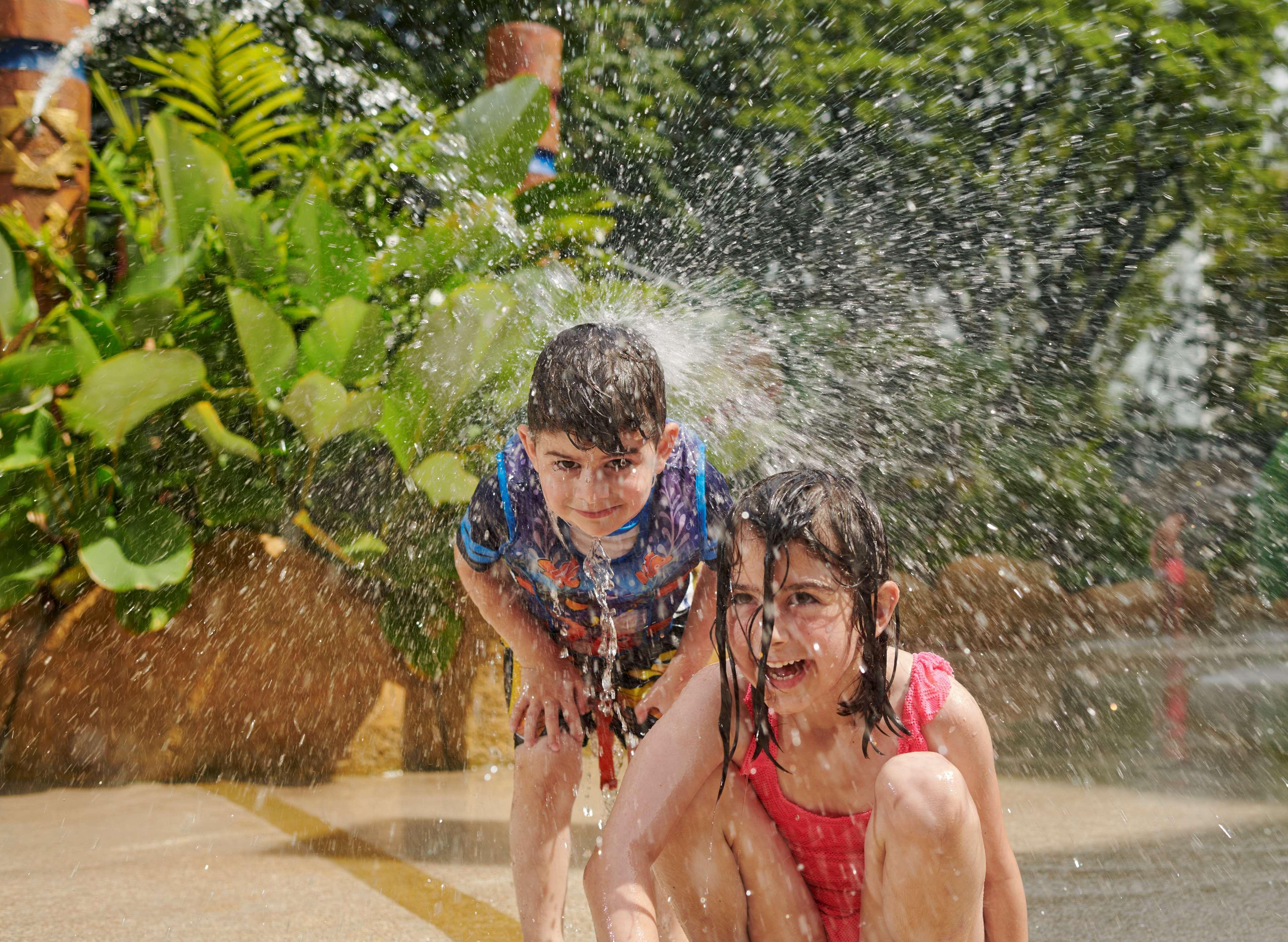 Parkroyal Penang Resort Batu Ferringhi Exterior photo The photo shows two children playing in water. One child, a boy, is standing and looks like he is getting splashed, while the other child, a girl, is crouching down, smiling, and also appears to be enjoying the water. They are surrounded by green pla