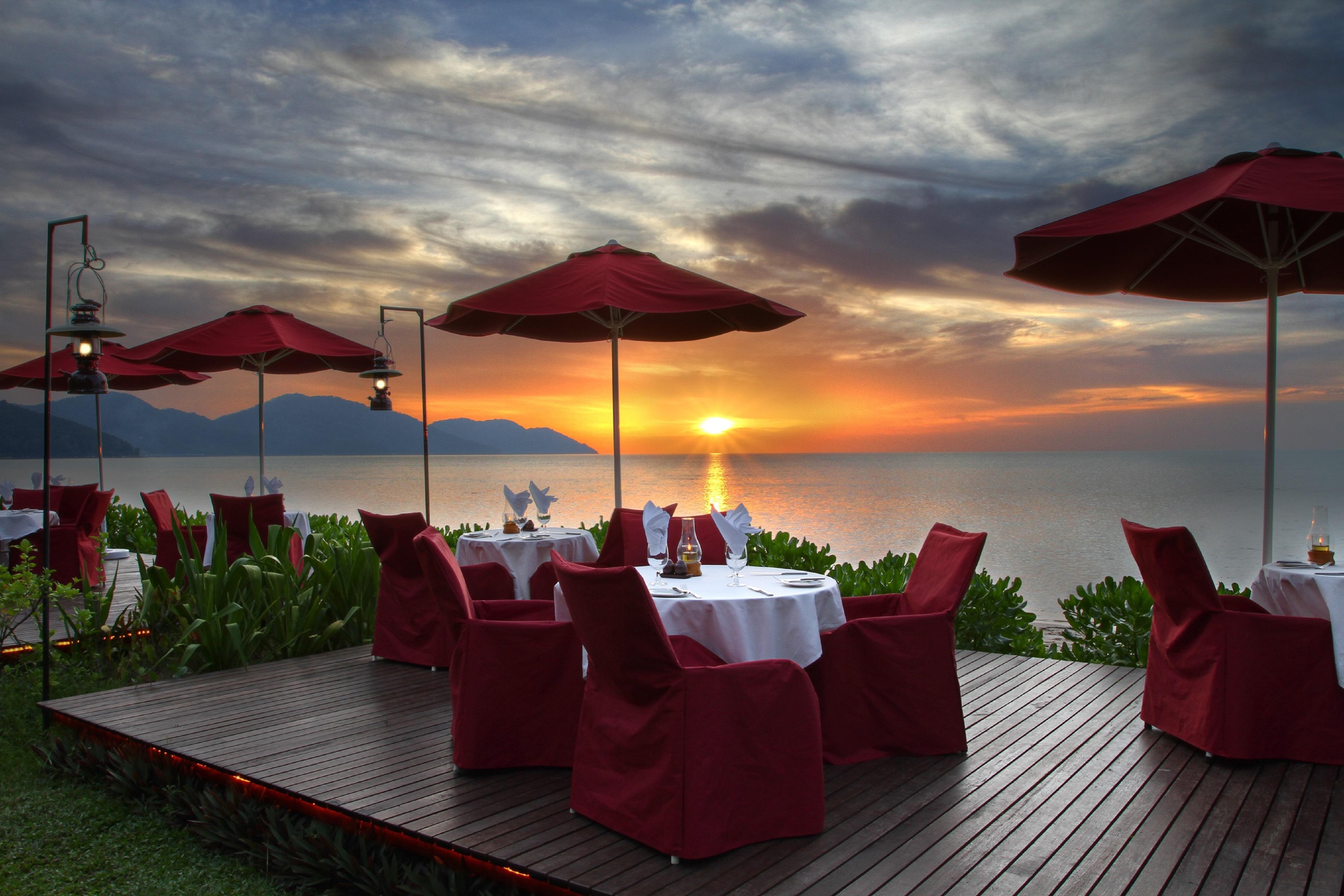 Parkroyal Penang Resort Batu Ferringhi Exterior photo The photo depicts a serene outdoor dining setting at a restaurant, with tables arranged on a wooden deck overlooking a body of water. Each table is elegantly set with white tablecloths and surrounded by red chairs, which add a pop of color. The scene