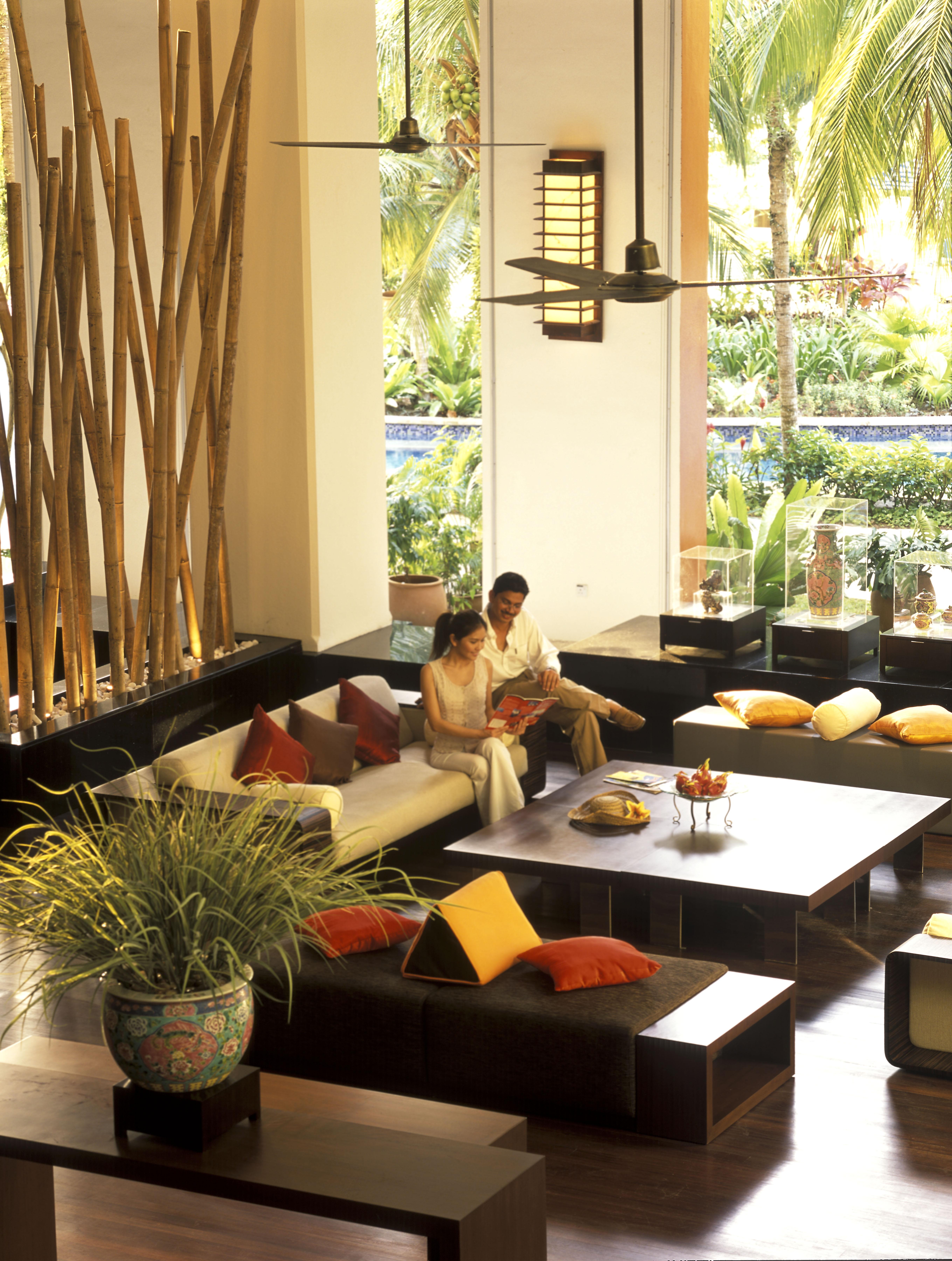 Parkroyal Penang Resort Batu Ferringhi Room photo The photo depicts a cozy and stylish lounge area. It features a couple seated on a plush white sofa, surrounded by colorful cushions in shades of orange and red. In the background, there are tall bamboo plants adding a natural touch. A coffee table i