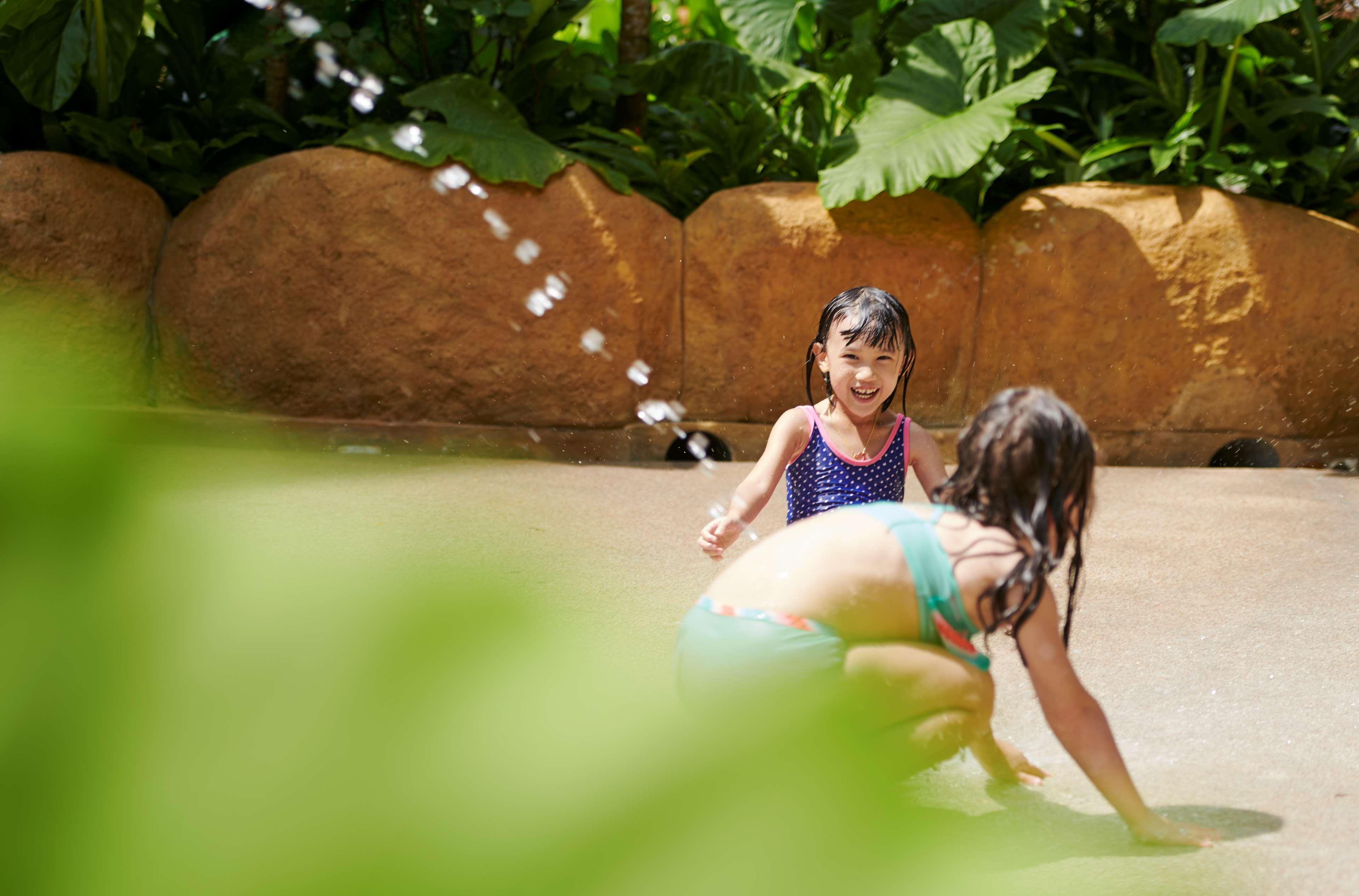 Parkroyal Penang Resort Batu Ferringhi Exterior photo The photo shows two children playing in a water area, possibly at a water park or pool. One child, wearing a colorful bikini, is kneeling and playing on the ground, while the other, in a blue swimsuit, is standing and looking happily at the camera. T