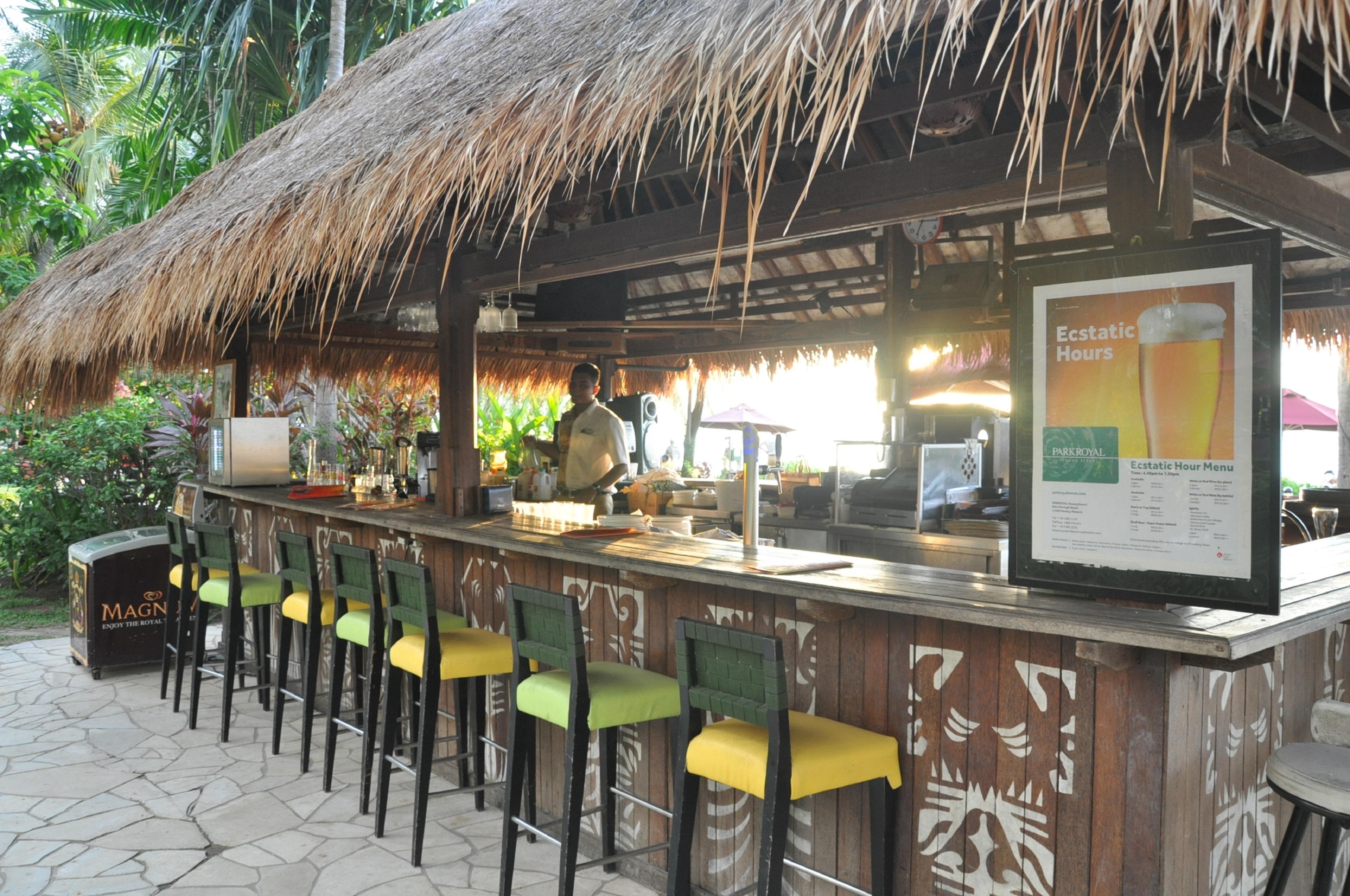 Parkroyal Penang Resort Batu Ferringhi Exterior photo The photo shows an outdoor bar with a thatched roof, characteristic of a tropical or resort setting. The bar is made of wood and features several bar stools with green and yellow seats. Behind the bar, a bartender is preparing drinks, with various in