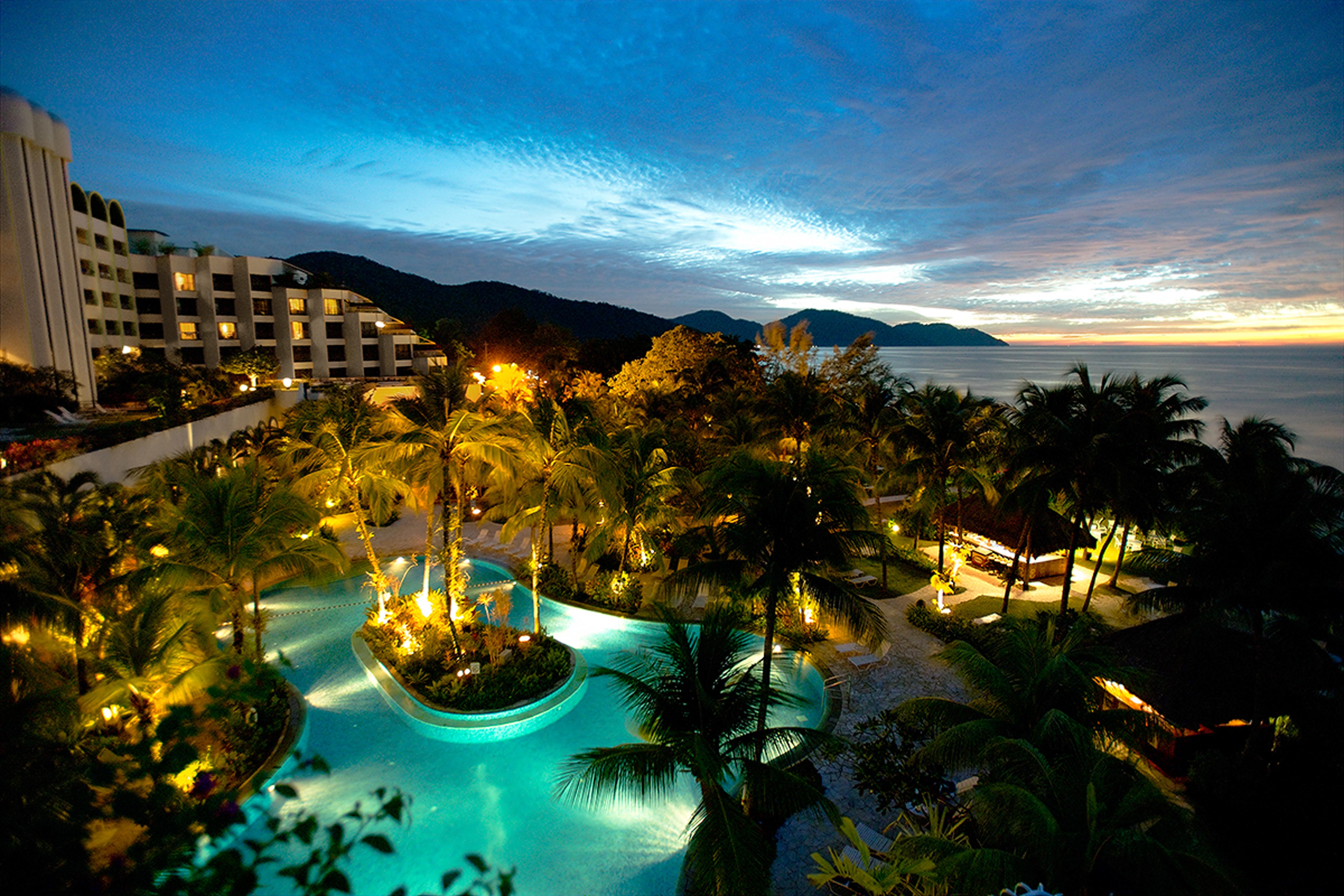 Parkroyal Penang Resort Batu Ferringhi Exterior photo The photo shows a scenic view of a resort area during dusk. In the foreground, there is a beautifully illuminated swimming pool surrounded by lush palm trees, creating a tropical atmosphere. The pool has a unique shape, and its water is glowing in a 