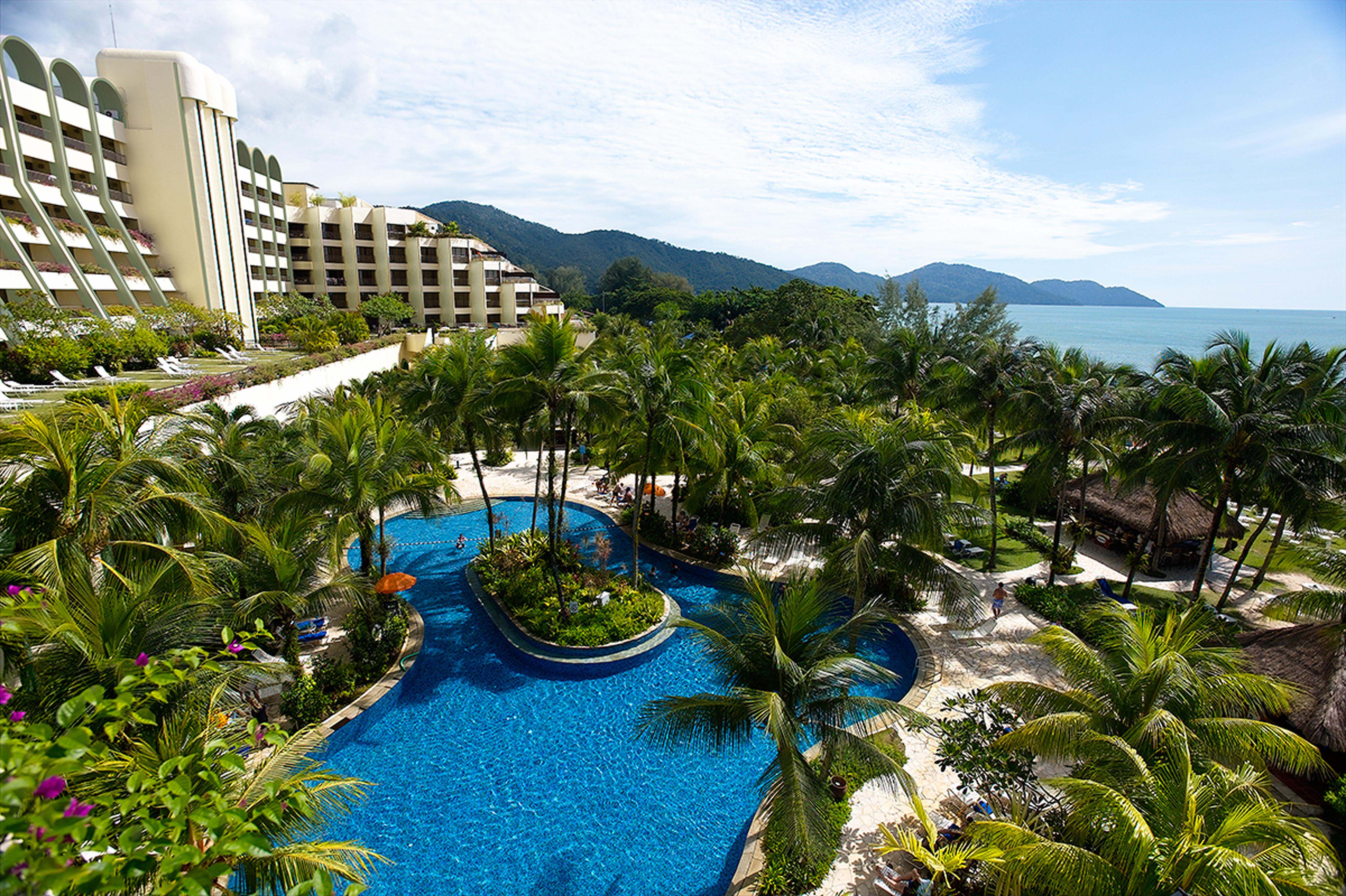 Parkroyal Penang Resort Batu Ferringhi Facilities photo The photo depicts a luxurious resort with several buildings, set amidst lush greenery and palm trees. In the foreground, there is a large, inviting swimming pool with a curved shape, surrounded by tropical plants and colorful flowers. Beyond the pool